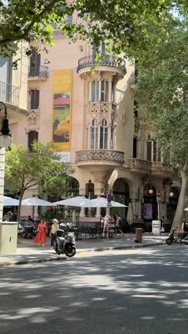 The video shows a street scene in front of a building with ornate architecture. There are several tables and umbrellas set up outside a cafe on the sidewalk, with a few people sitting at them. A scooter is parked on the sidewalk. A banner hangs from the building advertising an exhibition titled "COLORS DEL MON". Trees line the street, providing shade. A few other people are walking on the sidewalk and a motorcycle is parked further down the street. The overall impression is a sunny day in a European city.