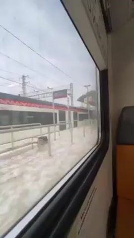 The video shows a view from inside a train, looking out of a window. Another train is parked on a snow-covered platform next to it. Above the parked train, there are power lines.