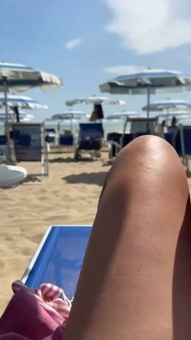 The video shows a person's bare legs and feet resting on a beach chair on a sunny day at the beach. The person is reclining on a blue beach chair with a pink and white striped towel draped over it. The background shows a sandy beach with rows of beach umbrellas and chairs. The sky is blue with a few clouds. The camera angle is from the perspective of the person, focusing on their legs and feet. The feet appear to have some sand on them.