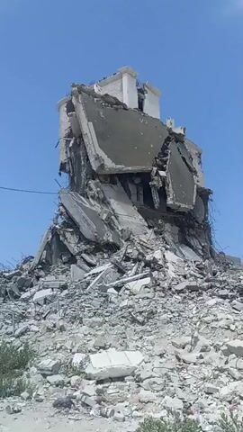 The video shows a partially collapsed building against a clear blue sky. The structure appears to be made of concrete and rebar, with large sections of walls and floors broken and fallen. Debris is piled high around the base of the building, suggesting significant destruction. There are no people visible in the footage. The environment appears to be an outdoor area, possibly urban or semi-urban, with some low-lying vegetation visible at the bottom of the frame.
