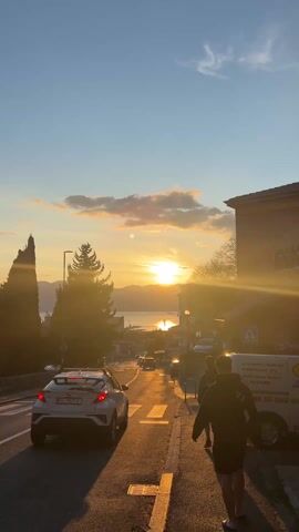 A street scene shows a sunset over the sea. There are some buildings on either side of the street. A couple of trees are also in view. A car is driving down the street, and some people are walking on the sidewalk. The sun is reflecting off the water. The sky is a clear blue with some clouds.