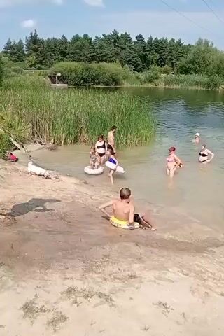The video shows a sunny day at a lake with a sandy shore and trees in the background. Several people are in the water, including adults and children, some of whom are wearing swimsuits. There are also inflatable rings and tubes in the water. A dog is visible on the shore. Some people are wading, while others are swimming or playing in the water. A child is splashing in the water, and another child is sitting on the shore. The overall atmosphere appears to be relaxed and recreational.