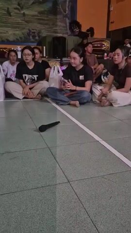 A group of young people, likely teenagers or young adults, are sitting on the floor in a large room with a tiled floor and yellow walls. There is a mural on the back wall. A microphone is on the floor in the foreground. The people are sitting in a semicircle, facing towards something or someone not fully visible in the frame. Some individuals are looking forward, while others are looking to the side or down. One person is holding a white plastic bag. In the background, there are some musical instruments, including drums. The lighting appears to be artificial, creating a slightly dim atmosphere. The overall impression is of a casual gathering or a rehearsal.