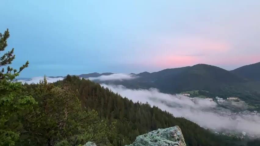 This is a video of a mountain landscape at dawn. The scene features rolling hills covered in lush green pine trees. A small village is nestled in the valley below, partially obscured by wisps of fog that drift through the trees. The sky transitions from a soft blue in the upper left to vibrant hues of pink and orange on the right, indicating the sun is beginning to rise. The overall atmosphere is serene and peaceful, with a sense of natural beauty and tranquility. There is no visible text in the video.