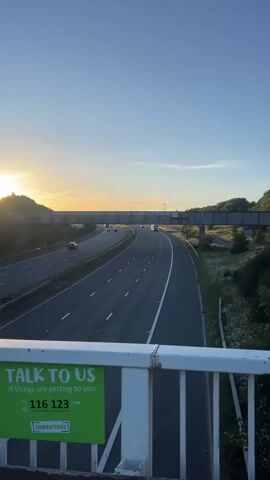 A vertical, static video captures a sunset over a highway. In the foreground, a white railing with a green sign is visible. The sign reads "TALK TO US If things are getting to you 116 123 Samaritans". The highway stretches into the distance, with cars and trucks driving on it. A pedestrian overpass crosses the highway. The sky is clear and blue, with the sun low on the horizon, casting a warm glow.