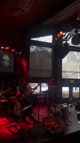 A band is performing on a stage in a bar or restaurant, bathed in red light. The band consists of a drummer, bassist, guitarist, and singer. The guitarist and singer are in the center of the stage, each standing at a microphone. The stage is cluttered with musical equipment. Behind the stage are large windows that show a snowy outdoor scene. Several patrons are seated at tables. A banner is visible in the background to the left. A disco ball hangs from the ceiling on the right.
