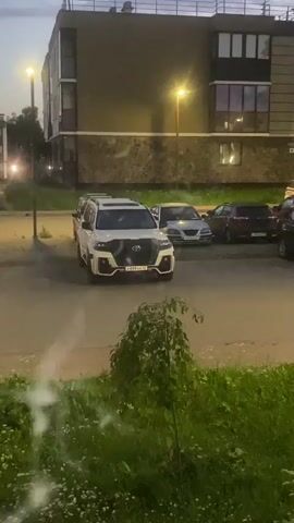 This is a brief video clip showing a parking lot outside a modern apartment building. The sky is dark, suggesting dusk or evening. Several cars are parked in the lot, including a prominent white SUV in the foreground. There is a significant amount of white smoke or vapor drifting across the scene, obscuring parts of the view and originating from an unseen source, possibly near the bushes in the lower foreground. The smoke appears to be dissipating into the air. The lighting in the scene comes from streetlights and interior lights from the building. No people are clearly visible.
