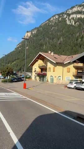 The video pans across a street in a mountainous area. On the right side of the street, there are several buildings, one of which is yellow and has balconies. A black utility truck with a trailer is parked on the side of the road. Further down the street, there are more buildings and trees. A large, forested mountain looms in the background. The sky is blue with some clouds. There are pedestrians, cars, and streetlights visible.