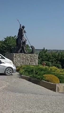 The video pans across a scenic overlook on a clear, sunny day. In the foreground, a portion of a statue, possibly depicting a figure fighting a dragon, is visible on a stone pedestal. Lush green bushes and manicured landscaping are arranged around the pedestal. The camera then moves to reveal a wrought-iron fence and a stone retaining wall. Beyond the fence, a sprawling town or city is visible in the valley, nestled among rolling hills. In the far distance, hazy mountains complete the picturesque landscape. The overall impression is of a well-maintained viewpoint offering a panoramic vista of a settlement and its natural surroundings.