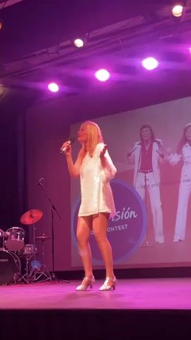 The video shows a woman singing on a stage. She is wearing a sparkly white dress and silver shoes. She is holding a microphone and gesturing with her hands. Behind her, a screen displays the Eurovision logo and images of ABBA. There is a drum set on the left side of the stage. The stage is lit with purple lights.