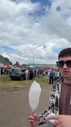 A medium shot shows a man in sunglasses and a plaid shirt holding a pink and white piece of cotton candy, biting into it. He is in the foreground, and in the background, a crowd of people are gathered at an outdoor event, possibly a fair or festival, under a cloudy sky. There are tents, cars, and a green hill in the distance. The man then turns to the camera and smiles as he continues to eat the cotton candy. The scene then shifts to show the crowd from a different angle, with more people walking around and interacting. The camera pans across the event, showing various stalls and activities, including a small white horse with a pink saddle. The video ends with a shot of people walking away from the camera, towards more tents and activity.