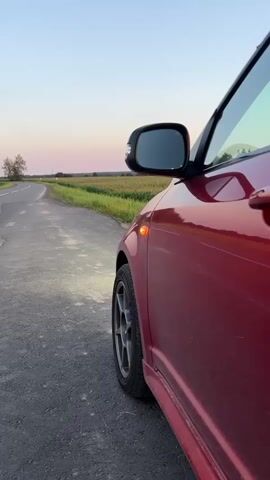 The video shows a side view of a red car parked on the side of a rural road. The car's turn signals blink. The road curves to the left in the distance, and the surrounding landscape consists of fields of tall grass or crops. The sky is clear with a gradient of colors suggesting either sunrise or sunset. The lighting is soft and ambient. There are no people visible in the video.
