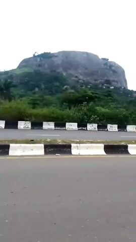 The video shows a roadside view with a large, rounded, rocky hill or small mountain in the background. The hill is covered in greenery at its base and has some trees on top. In the foreground is a paved road with a black and white striped barrier along the edge. A thin lamppost is also visible along the roadside. The sky is overcast. The camera appears to be moving.