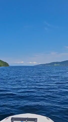 The video shows a view from a boat on a blue body of water under a clear blue sky with a few scattered clouds. The water has moderate ripples. In the distance, a green, tree-covered hill slopes down to the water's edge. There are some buildings visible on the hillside. The front of a white boat is visible in the lower portion of the frame. The video is taken from a moving perspective, suggesting the boat is underway.