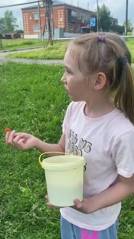 A young girl with blonde pigtails and a pink t-shirt stands in a grassy area, holding a yellow bucket in her left hand. She is looking to her right, and with her right hand, she brings a small red berry to her mouth and eats it. The bucket contains several red berries. In the background, there is a brick building, a street, and trees.