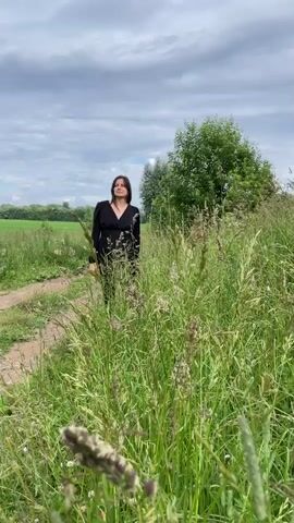 A young woman with dark hair stands in a field of tall green grass. She wears a long-sleeved black dress. The sky above is cloudy and grey. She initially stands with her arms down, then extends them outwards, and finally raises them above her head.