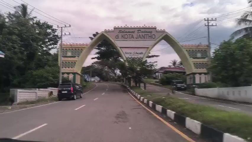 The video shows a car driving on a road through a city gate in Jantho, Indonesia. The gate is made of concrete and has two arches with a sign that reads "Selamat Datang di Kota Jantho" ("Welcome to Jantho City"). The gate is decorated with traditional Islamic patterns. There are trees and buildings on either side of the road. There are two cars and one person on a bicycle visible in the distance. The sky is overcast.