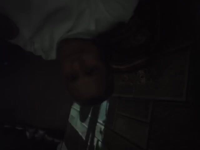A dimly lit video shows a man in a white t-shirt sitting or reclining. He is facing the camera with a neutral expression. Behind him is a patterned pillow and a wall with a textured surface. A window or opening casts a vertical shadow on the wall behind him. Additional items are indistinctly visible on a shelf in the upper right corner.