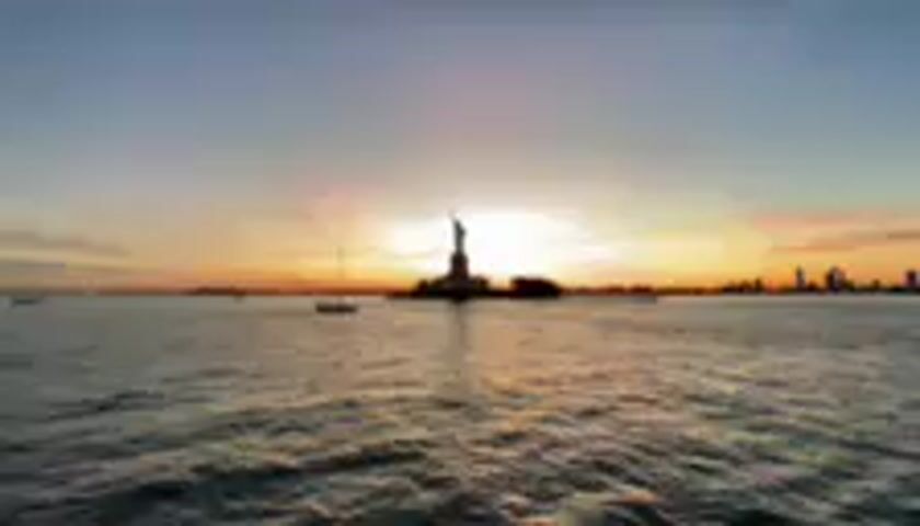 The media file captures a serene sunset over the water, with the iconic Statue of Liberty silhouetted against the vibrant sky. The sun is setting behind the statue, casting a warm glow on the horizon. The water in the foreground is slightly choppy, reflecting the colors of the sunset. In the distance, the New York City skyline is faintly visible on the right. A few small boats are scattered across the water, adding to the peaceful atmosphere. The sky transitions from a deep blue at the top to shades of orange, pink, and yellow near the horizon. The overall mood is tranquil and awe-inspiring, evoking a sense of wonder and patriotism. The time of day is clearly sunset, and the location appears to be a body of water offering a clear view of the Statue of Liberty and the Manhattan skyline, consistent with views from New Jersey. There is no visible text in the media file.