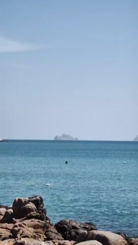 The video shows a scenic view of the ocean on a clear, sunny day. In the distance, there are two islands, one with a distinctive pyramid-like shape. In the foreground, there is a rocky shoreline. The water is a bright, clear blue. The camera slowly zooms in on the pyramid-shaped island.