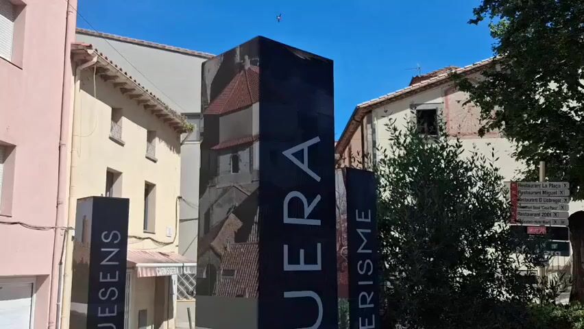 The video shows a street in the town of Requesens in Spain. The street is lined with buildings, including a pink building on the left. There are three black rectangular signs with text on them. The signs advertise tourism in the area. The text on the signs includes "REQUESENS", "CONQUERISME", "LA JONQUERA", and "SENDERISME". There is a blue car parked nearby. The sky is blue and there are some trees visible. The video then pans down to show the base of the signs.