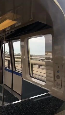 The view is from inside a train or monorail as it travels along tracks. The camera is positioned near a window, and the focus is on the exterior view. Through the window, there is a scene of an airport runway or airfield, with the edge of a building visible in the distance. The sky is partly cloudy with dark, ominous clouds appearing to the right of the frame. The interior of the train includes a dark patterned floor, off-white walls, and a small panel with buttons and a speakerphone. The carriage is likely moving.