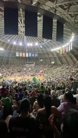 The video shows a large crowd of people inside a stadium. The stadium has a domed ceiling with lights and banners hanging from it. In the center of the stadium is a basketball court, where a game appears to be in progress. Many people in the crowd are wearing green shirts and hats, and some are waving flags. The atmosphere appears to be lively and exciting.