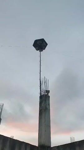 A tall concrete pillar stands in the center of the frame, topped with exposed rebar and what appears to be a small electrical box. Attached to the pillar is a long, slender pole, also made of metal, that extends upwards to support a large, hexagonal object. This object has multiple speakers or lights pointing outwards in different directions. Thin wires extend from the top of the pillar and the hexagonal device, stretching towards the edges of the frame, possibly acting as guy wires for stabilization. A low concrete wall forms the bottom of the frame. The sky above is overcast, with subtle hints of pink and orange near the horizon, suggesting either sunrise or sunset. The overall scene suggests an outdoor utility structure, possibly for public address or lighting, on a cloudy day.