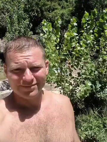 The video begins with a close-up of a shirtless man looking at the camera with a slight smile. He is in the foreground, and the background is out of focus. As the video progresses, the camera pans slightly to the right, revealing a body of clear turquoise water with lush green foliage and trees surrounding it. Several people are visible in the water and on the rocky banks, some swimming, others sitting or standing. The overall environment appears to be a natural, outdoor setting, likely a river or natural pool. The man in the foreground remains the primary focus throughout the short clip.