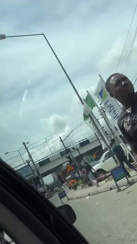 The video shows a street scene in what appears to be a city in Nigeria. The view is from inside a car, looking out at the street. There's an overpass in the background, supported by pillars. A couple of pedestrians are walking on the sidewalk. Several "keke napep" (also known as auto rickshaws or tuk-tuks) are moving on the road. One is yellow and looks older, while another is blue and appears newer. A building with a large red logo is visible on the right side of the frame. Utility poles and a lot of wires run above the street. The sky is partly cloudy. A Nigerian flag is seen flapping in the wind.