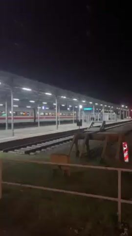 The video shows an exterior shot of a train station at night. The sky is dark, and the area is lit by the bright lights of the station. A white and red train is parked along the platform. A man wearing a dark jacket and hood walks near the edge of the platform. The ground is covered in grass and gravel.