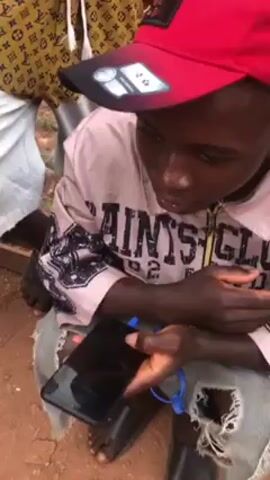 A young man wearing a pink shirt with a red baseball cap, ripped jeans, and black sandals sits outside, holding a black smartphone. The phone is connected to blue earphones. He appears to be engaged in a conversation, gesturing with his hands while looking at the phone. Another person is partially visible standing behind him, wearing a patterned shirt and light-colored pants. The ground appears to be reddish dirt or soil.