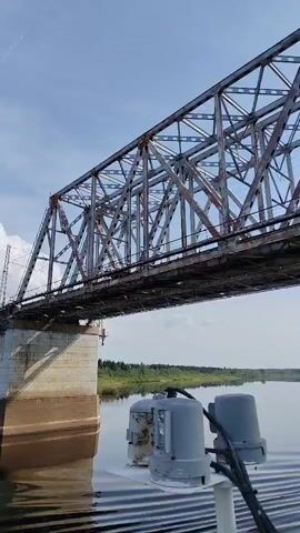 The video shows a steel truss bridge from a low vantage point on a boat passing beneath it. The bridge, supported by concrete pillars in the water, stretches horizontally above the river. Its steel framework appears slightly rusted. The water reflects the bridge and the sky. Two gray metal objects with black cables sit on the boat's surface. A green shoreline with trees runs along the distant riverbank. The sky is blue with scattered clouds. The boat is moving and a steady droning sound is audible.