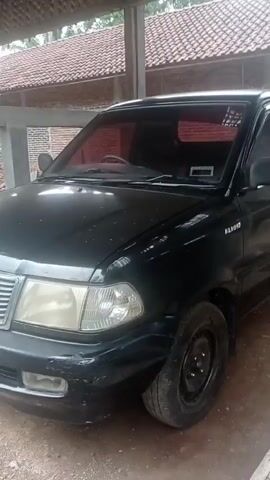 The video shows a black Toyota Kijang pickup truck parked under a simple, open-sided structure with a corrugated roof. The truck is facing forward, with its front end clearly visible. The paint on the truck appears worn and scratched, particularly on the front bumper. The headlights are visible, as is the grille with the Toyota logo. The license plate, partially obscured, reads "8446 BL". The interior of the truck has tinted windows with a reddish hue. In the background, there is a brick wall and some roofing tiles. To the left of the truck, a wheelbarrow with yellow containers and some wooden planks are visible. The ground is dirt. The overall impression is that of a vehicle in a rural or semi-rural setting, possibly undergoing some form of repair or storage. There are no people visible in the video.