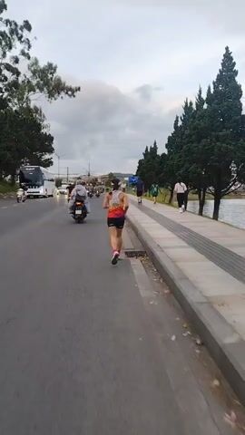 The video shows a person running on a sidewalk next to a road. The runner is wearing a white tank top with colorful stripes and black shorts. They are also wearing a black visor and pink and white running shoes. The road is paved and has multiple lanes of traffic. Several motorcycles and cars are visible on the road. On the other side of the sidewalk is a body of water, possibly a lake, and a line of trees. Other people can be seen walking and running in the distance. The sky is cloudy and overcast. The video appears to be filmed from a moving perspective, likely following the runner.