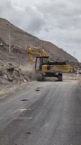 A bright yellow excavator is seen digging into a rocky hillside, spewing dust into the air. The excavator is on the right side of a paved road, and is working to clear debris from the side of the road. In the background, there are other construction vehicles and a mountainous landscape under a cloudy sky. The road appears to be in disrepair, with visible potholes.