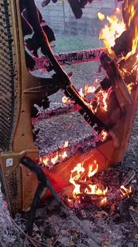 A piano is burning outdoors. The piano is upright and appears to be partially disassembled. Flames are visible inside and outside of the piano. The piano is on the ground in an outdoor setting with dry leaves and debris around it.