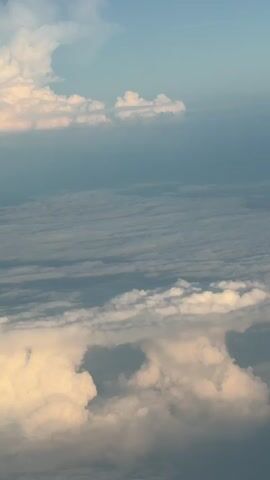 The video shows a view from an airplane window, looking out at clouds and the sky. The clouds are white and fluffy, with some appearing to be illuminated by the sun. The sky is blue, with some wispy clouds. The wing and engine of the airplane are visible in the foreground. The video appears to be filmed during the daytime, likely in the morning or evening, given the soft lighting.