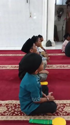 The video shows a group of young boys sitting on a red carpet inside a mosque. They are all wearing traditional Muslim attire, including kufis, and appear to be listening to an older person who is speaking to them. The mosque has white walls and pillars, and there is a wooden pulpit in the background. The lighting is dim, and the overall atmosphere seems to be one of reverence and learning.