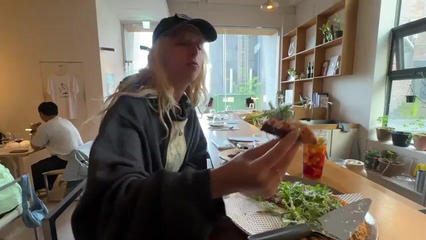 A person with long blonde hair, wearing a black cap and a dark sweatshirt, is standing at a wooden counter in a bright cafe or restaurant. They are holding up a piece of food, topped with green sprouts, and taking a large bite. In front of them is a plate with more of the same food, accompanied by a serving utensil. A glass of iced tea with a lemon wedge is on the counter as well.  A man is seated at a table in the background, and a t-shirt is hung on the wall. The restaurant has a window and shelves containing books and plants.