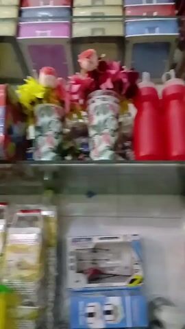 The video shows the interior of a store, likely a discount or dollar store, with shelves stocked with various household items. The shelves contain items such as plastic containers, decorations, kitchenware, and other assorted goods. Several customers, some wearing head coverings, are browsing the merchandise. There is at least one sign that reads "$1" indicating a price point. The lighting in the store is somewhat bright, and the store appears relatively crowded with merchandise.