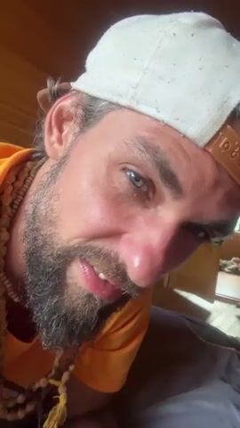 The video features a close-up, low-angle shot of a man with a beard, wearing a white baseball cap and an orange shirt. He has necklaces made of beads around his neck. The man is looking directly at the camera with a serious expression. The background appears to be a dimly lit room with wooden walls, possibly a cabin or a rural dwelling. The lighting suggests it could be daytime, but the indoor setting makes it difficult to be certain. The man's face is illuminated, highlighting his features and the texture of his beard and skin. The activity appears to be the man posing or interacting directly with the camera. There is no visible text in the video. The overall mood is intense and personal due to the close proximity and direct gaze.