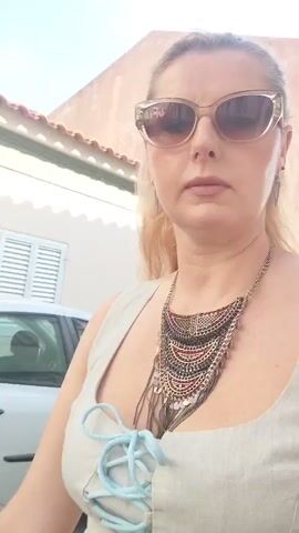 A woman with blonde hair and sunglasses is standing outdoors and filming herself with a phone. She is wearing a light-colored sleeveless top with a light blue lace-up front and a large, ornate necklace. In the background, there are cars parked on a cobblestone street. A white car is visible on the left side of the frame, and a dark-colored car is in the distance on the right. The woman is looking at the camera and appears to be moving forward.