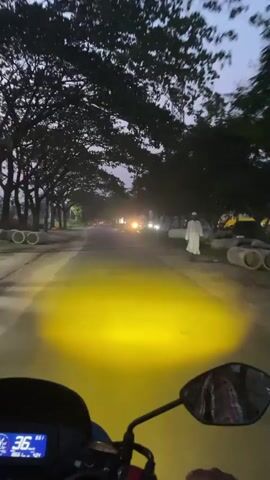 This video captures a first-person perspective from a motorcycle riding along a tree-lined road in Nasirabad, Bangladesh, during dusk or twilight. The sky is a muted grey-blue, indicating the transition from day to night, with vehicle headlights already switched on. A prominent yellow beam from the motorcycle's own headlight illuminates the paved road ahead.

On the right side of the road, a person dressed in a long white robe, possibly a thobe or jubbah, is seen walking calmly in the same direction as the motorcycle. Further along the right side, large concrete pipes are stacked, suggesting ongoing construction or material storage. Both sides of the road are densely bordered by mature trees, their dark silhouettes contrasting against the lighter sky. Utility poles or transmission towers are also visible in the distance through the gaps in the trees.

The dashboard of the motorcycle is visible at the bottom of the frame, showing a digital speedometer. The numbers "36" and "65.1" are discernible, likely indicating a speed of 36 km/h and a trip or odometer reading of 65.1 km. Other vehicles with illuminated headlights are seen approaching in the opposite lane, creating points of light in the receding distance. The scene conveys a sense of peaceful transit through a semi-urban or rural environment as daylight fades.