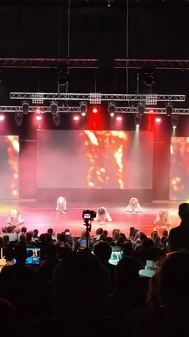 A stage performance is captured from the audience perspective. Seven dancers, predominantly female, are on a brightly lit stage, dressed in what appear to be revealing costumes. The stage features a large screen displaying abstract, fiery visuals. The dancers are engaged in synchronized movements, including low stances and dynamic poses. Smoke or fog is present on stage, adding to the dramatic lighting effects. In the foreground, the silhouettes of audience members are visible, some holding up phones to record the performance. A camera on a tripod is positioned between the audience and the stage. The overall atmosphere suggests a contemporary dance or performance art piece.