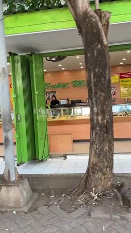A street-level view shows the exterior of a shop with a bright green accordion gate partially open. Inside, a woman wearing a headscarf stands behind a counter in a well-lit store. Display cases filled with items are visible. The shop's signage, "Bukit Mas," is illuminated. A tree with a thick trunk stands prominently in the foreground, partially obscuring the view. The ground is paved with bricks, and there is debris scattered around the base of the tree and sidewalk. The overall environment appears to be an urban or semi-urban street setting.