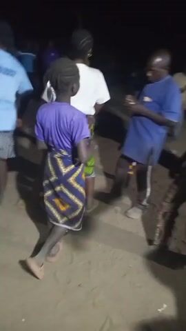 The video captures a group of people dancing in an outdoor setting at night. The area is sandy and dimly lit, with several individuals visible in the background. The focal point is a group of three people. A young person in a purple t-shirt and patterned wrap stands with their back to the camera, seemingly observing the dance. To their left, a woman in a white shirt and green patterned wrap dances briefly before exiting the frame. To the right, a woman in a red shirt and patterned pants energetically dances and moves her hips, enjoying the music. The sound of what appears to be amplified percussion and singing can be heard.