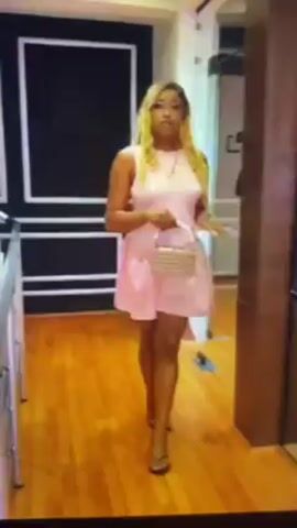 A woman with blonde hair and a light pink dress walks across a wooden floor. She carries a small, light-colored purse. The woman is indoors, with a door and a display case visible in the background. She turns slightly to the side, showing the back of her dress, which has ribbons.