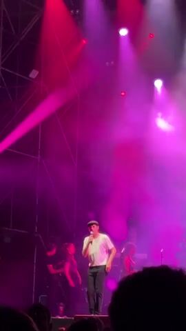 A male singer, dressed in a white t-shirt, black pants, and a black cap, sings into a microphone on a stage bathed in pink light and fog. He is gesturing with his hands as he performs. Behind him are musicians playing instruments: a guitarist on the left, a keyboardist in the center, and two backing vocalists on the right. The stage is backed by a metal structure. The audience is visible at the bottom of the frame.
