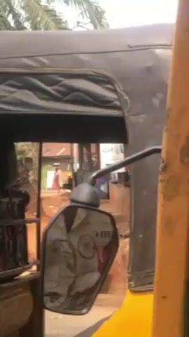 The video is shot from inside a yellow auto rickshaw, commonly known as a "Keke" or "Tuk-Tuk," moving along a street. The focus is on the view through the open sides of the vehicle, showcasing a street scene with various structures and people. In the background, palm trees and small shops with signs are visible. A man wearing a red tank top can be seen standing near one of the shops. The camera captures the driver's hands as they adjust a mirror, suggesting they are preparing to continue the journey. The overall setting indicates a tropical or subtropical environment, likely a small town or village. The street surface appears to be unpaved and possibly muddy.